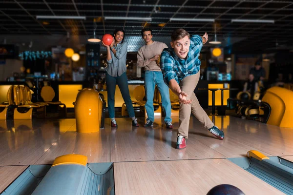 Sonriente joven lanzando bowling bowl en skittle alley cerca de amigos multiculturales - foto de stock