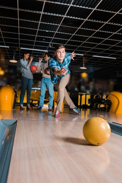 Konzentrierter junger Mann wirft Bowlingschüssel auf Kegelbahn bei multikulturellen Freunden — Stockfoto