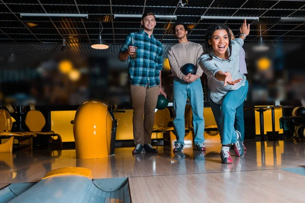 Joyeuse fille afro-américaine jetant bol de bowling sur ruelle de skittle près d'amis multiculturels — Photo de stock