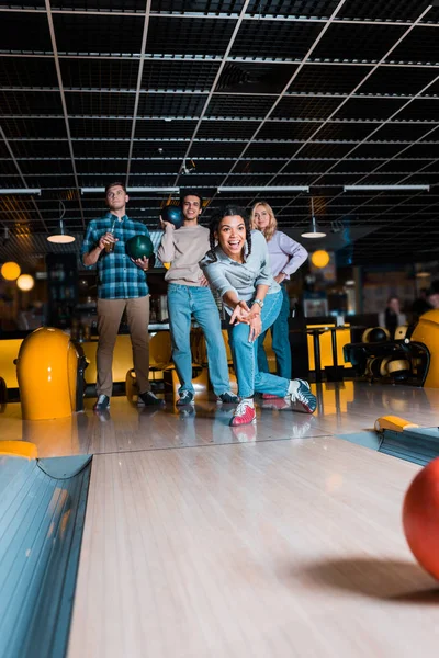 Eccitato africano americano ragazza lancio palla da bowling su skittle vicolo vicino a amici multiculturali — Foto stock