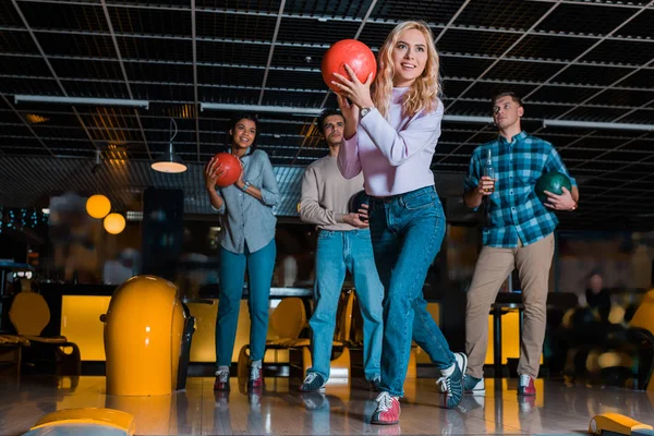 Smiling blonde girl throwing bowling ball near multicultural friends — Stock Photo