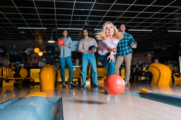 Sonriente chica rubia lanzando bola de bolos en el callejón de skittle cerca de amigos multiculturales - foto de stock