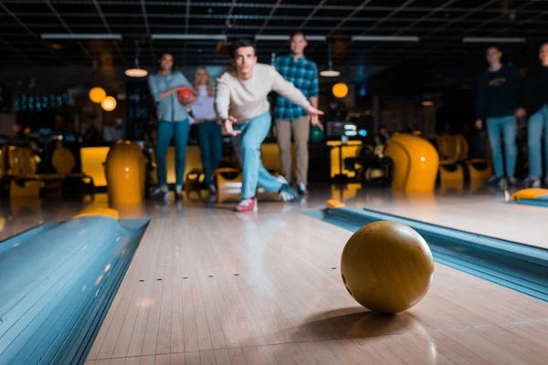 Selective focus of young man throwing bowling ball on skittle alley near multicultural friends — Stock Photo