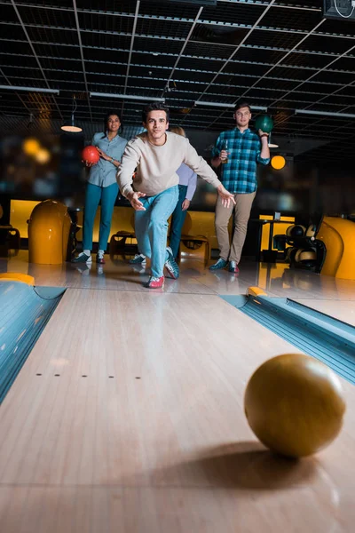 Handsome young man throwing bowling ball on skittle alley near multicultural friends — Stock Photo