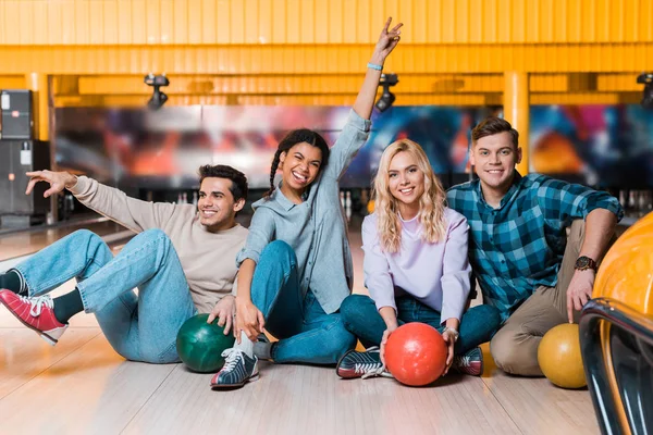 Happy african american girl showing victory gesture while sitting with multicultural friends on skittle alley in bowling club — Stock Photo