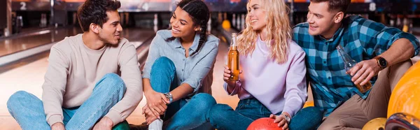 Panoramic shot of happy multicultural friends with bottles of beer sitting and talking on skittle alley in bowling club — Stock Photo