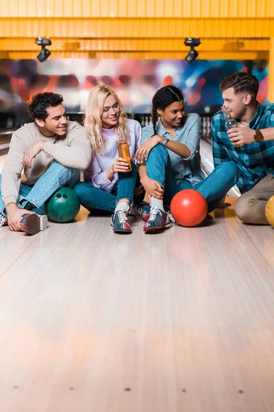 Cheerful multicultural friends with bottles of beer sitting and talking on skittle alley in bowling club — Stock Photo