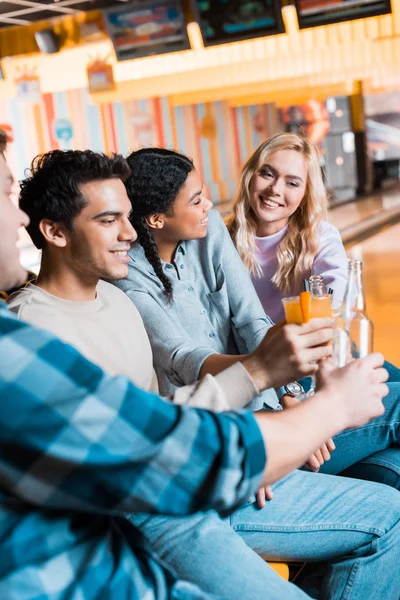 Amici multiculturali sorridenti che tengono bottiglie di birra e bicchieri da cocktail mentre sono seduti nel bowling club — Foto stock