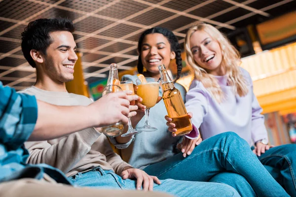 Cheerful multicultural friends clinking with bottles of beer and cocktail glasses while sitting in bowling club — Stock Photo