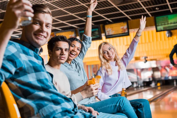 Amigos multiculturales felices sonriendo a la cámara mientras se sienta en el club de bolos con cócteles y cerveza - foto de stock