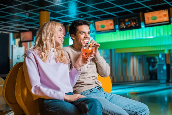 Alegre pareja mirando hacia otro lado y sonriendo mientras bebe cócteles en el club de bolos - foto de stock