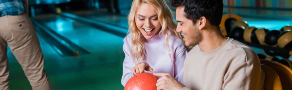 Панорамный снимок веселой пары, смотрящей на мяч для боулинга, сидящей в боулинг-клубе — стоковое фото