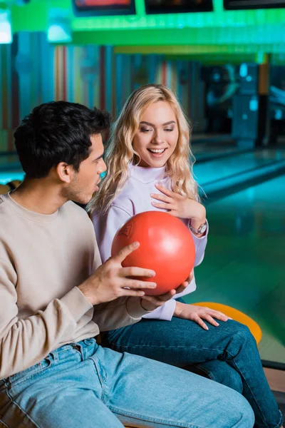 Surpresa, sorrindo menina tocando no peito enquanto namorado mostrando bola de boliche — Fotografia de Stock
