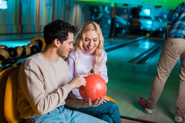 Hombre y mujer alegre mirando la bola bowilng mientras está sentado en el club de bolos - foto de stock