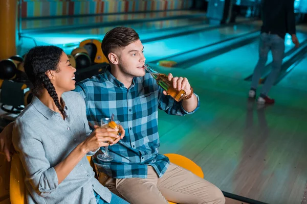 Alegre afroamericana chica celebración cóctel vaso y hablando con novio beber cerveza en bowling club - foto de stock