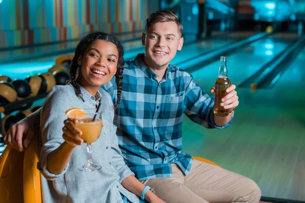 Feliz casal interracial olhando embora enquanto sentado com coquetel e cerveja no clube de boliche — Fotografia de Stock