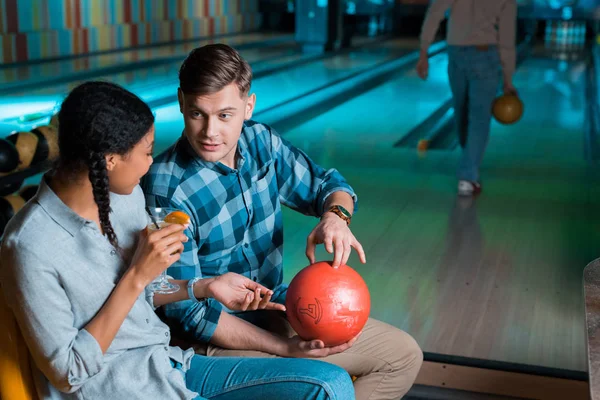 Handsome boyfriend showing bowling ball to cheerful african american girl holding glass of cocktail — Stock Photo