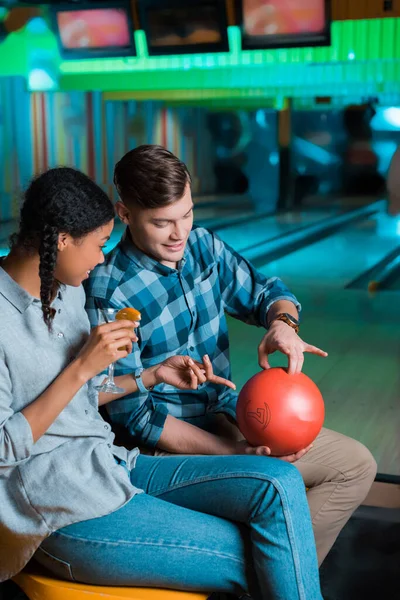 Sonriente novio mostrando bola de boliche a alegre africana americana chica sosteniendo vaso de cóctel - foto de stock