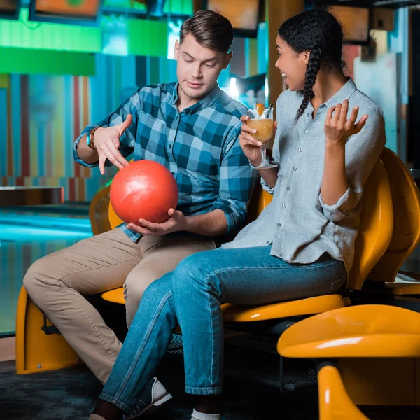 Sonriente afroamericano chica sosteniendo vaso de cóctel mientras novio mostrando bola de bolos - foto de stock