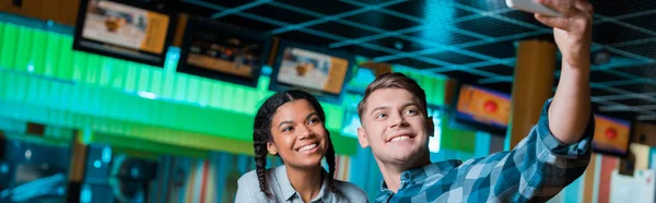Panoramic shot of happy interracial couple taking selfie on smartphone while sitting in bowling club — Stock Photo