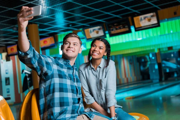 Улыбающаяся межрасовая пара делает селфи на смартфоне, сидя в боулинг-клубе — стоковое фото