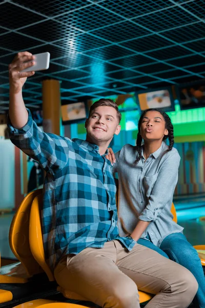 Alegre interracial pareja tomando selfie en smartphone mientras sentado en bowling club - foto de stock