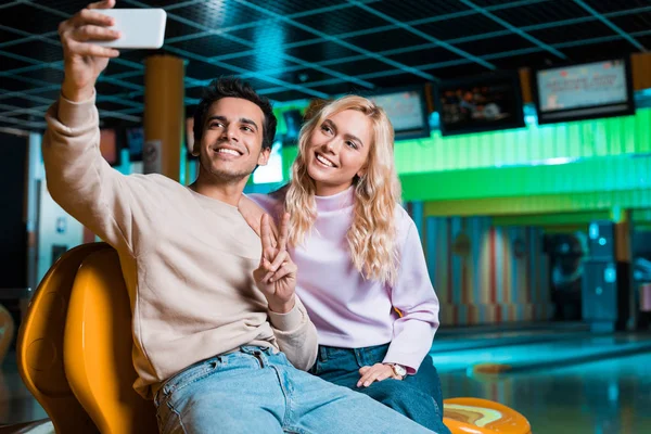 Happy young man showing victory gesture while sitting in bowilng club and taking selfie with smiling girlfriend — Stock Photo