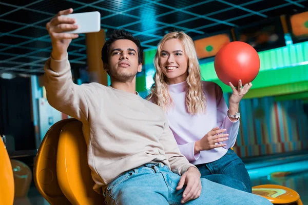 Happy girl holding bowling ball while boyfriend taking selfie on smartphone — Stock Photo