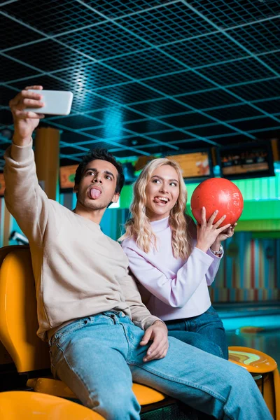 Веселая пара, торчащая языками и делающая селфи на смартфоне, пока девушка держит шар для боулинга — стоковое фото