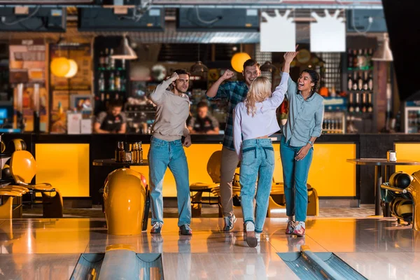Happy multicultural friends showing winner gestures near skittle alley in bowling club — Stock Photo