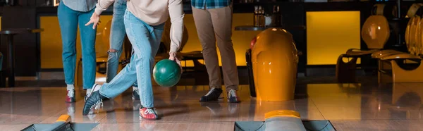 Cropped view of man throwing bowling ball on skittle alley near multicultural friends, panoramic shot — Stock Photo