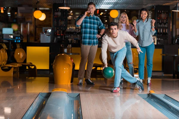 Schöner junger Mann wirft Bowlingball auf Kegelbahn in der Nähe multikultureller Freunde — Stockfoto