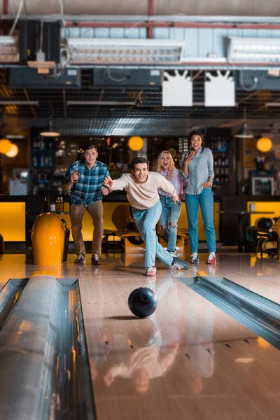 Handsome young man throwing bowling ball on skittle alley near multicultural friends — Stock Photo