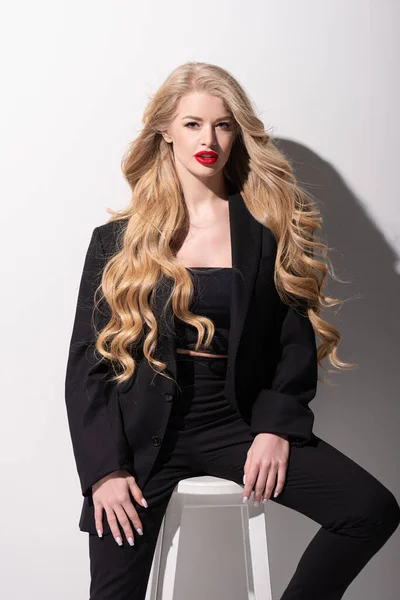 Curly young woman with red lips sitting on chair and looking at camera on white — Stock Photo