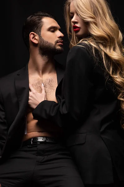 Young woman touching sexy man in suit sitting on chair isolated on black — Stock Photo