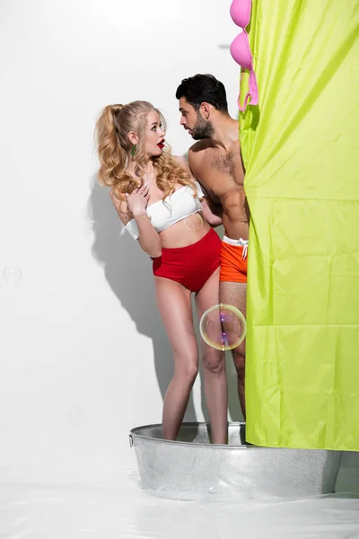 Impactado pin up mujer y hombre barbudo mirándose unos a otros cerca de la cortina de ducha en blanco - foto de stock