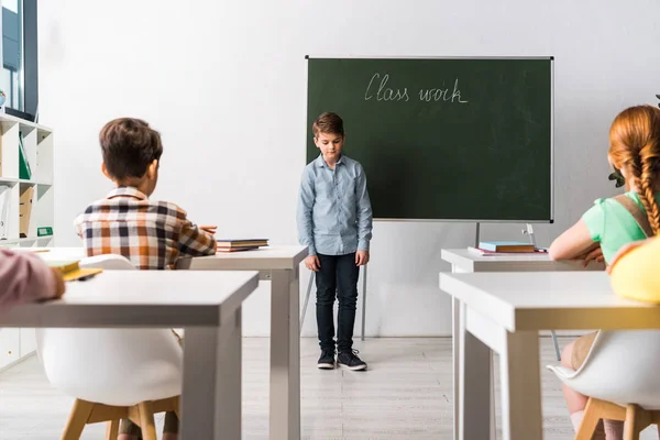 Schoolkids sitting at desks near classmate near chalkboard with class work lettering — Stock Photo