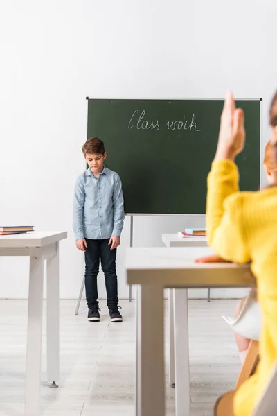 Foyer sélectif de l'écolier bouleversé debout près de tableau noir et camarade de classe avec la main levée — Photo de stock