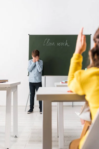 Selective focus of upset schoolboy covering face while standing near chalkboard and classmate with raised hand — Stock Photo