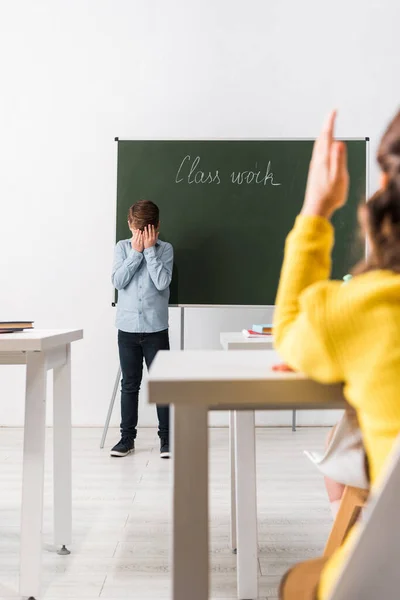 Foyer sélectif de l'écolier bouleversé couvrant le visage tout en se tenant près de tableau noir et camarade de classe avec la main levée — Photo de stock