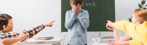 Colpo panoramico di compagni di classe che puntano con le dita al viso coperto da scolaro frustrato, concetto di bullismo — Foto stock