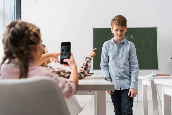 Selective focus of schoolboy pointing with finger at classmate while schoolgirl taking photo, cyberbullying concept — Stock Photo