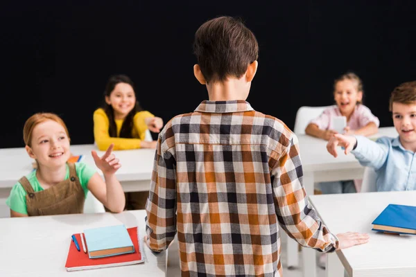 Back view of schoolboy standing near schoolkids pointing with fingers  isolated on black, bullying concept — Stock Photo