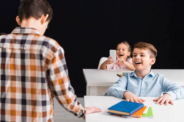 Enfoque selectivo de la colegiala tomando fotos de su compañero de clase, mientras que el escolar riendo aislado en negro, concepto de acoso cibernético - foto de stock