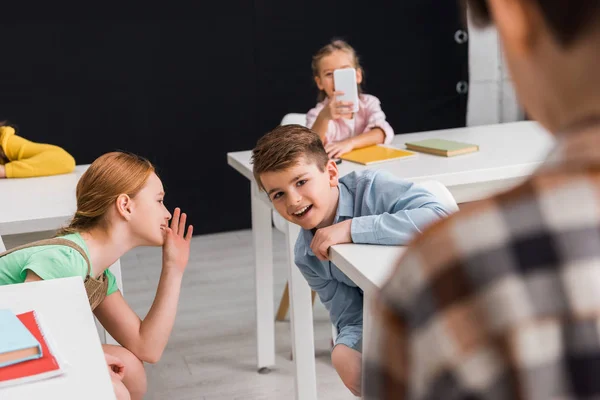 Selektiver Fokus von Schulkindern, die tratschen, während Schulmädchen Fotos von Klassenkameraden über schwarzes Cybermobbing-Konzept machen — Stockfoto