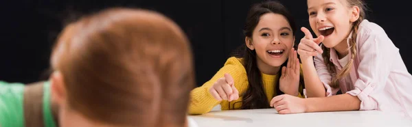 Панорамний знімок щасливих школярів, що вказують пальцями в однокласниках ізольовані на чорному, концепція знущань — стокове фото