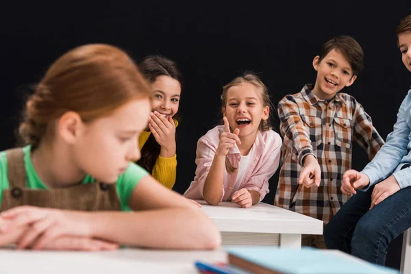 Enfoque selectivo de los compañeros de clase riendo y señalando con los dedos a la colegiala aislado en negro, concepto de intimidación - foto de stock