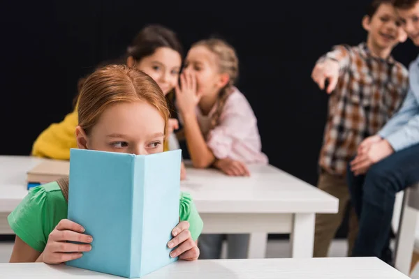 Messa a fuoco selettiva di sconvolto scolaro che copre il viso con libro vicino compagni di classe pettegolezzi e puntando con il dito isolato sul nero, concetto di bullismo — Foto stock