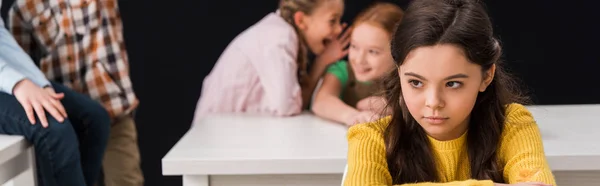 Panoramic shot of upset schoolkid near classmates gossiping and laughing isolated on black, bullying concept — Stock Photo