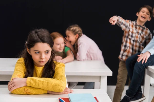 Enfoque selectivo de la colegiala molesta cerca de compañeros de clase chismorreando mientras el colegial señala con el dedo aislado en negro, concepto de intimidación — Stock Photo